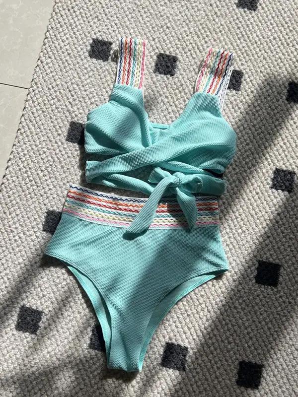 Aqua swimsuit with rainbow ribbon details on a patterned rug